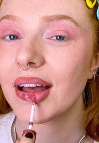 Applikator for flytende leppestift holdes nær leppene, påfører en blank nude nyanse. Har en slank, spiss applikator og en rosa-brun farge.