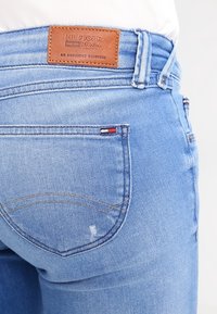 Ljust blå denimjeans med en läderetikett som har texten "HILFIGER DENIM", bakficka med subtila röda och vita sömnadsdetaljer.