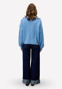Blauer, flauschiger Pullover mit tiefen Schultern, kombiniert mit weiten, dunklen Jeans aus Denim, die an den Säumen heller sind und Rückentaschen mit Akzenten aufweisen.