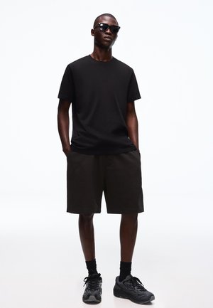 Mężczyzna w czarnych okularach przeciwsłonecznych, zwykłej czarnej koszulce, czarnych szortach, czarnych skarpetkach i czarnych trampkach stoi z rękami w kieszeniach.