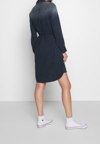 Abito camicia blu navy con maniche lunghe, vita cinte e orlo arrotondato, abbinato a sneakers bianche alte. Tessuto morbido, design casual.