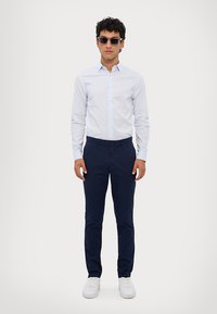 Lyseblå langermet skjorte, marineblå bukser, og hvite sneakers. Skjorten har en knapp-down design; briller er inkludert i antrekket.