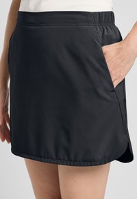 Zwarte sportieve rok met een elastische tailleband, zijzakken en een gladde, lichte stof met een subtiele glans en een schuin afgebroken zoom.