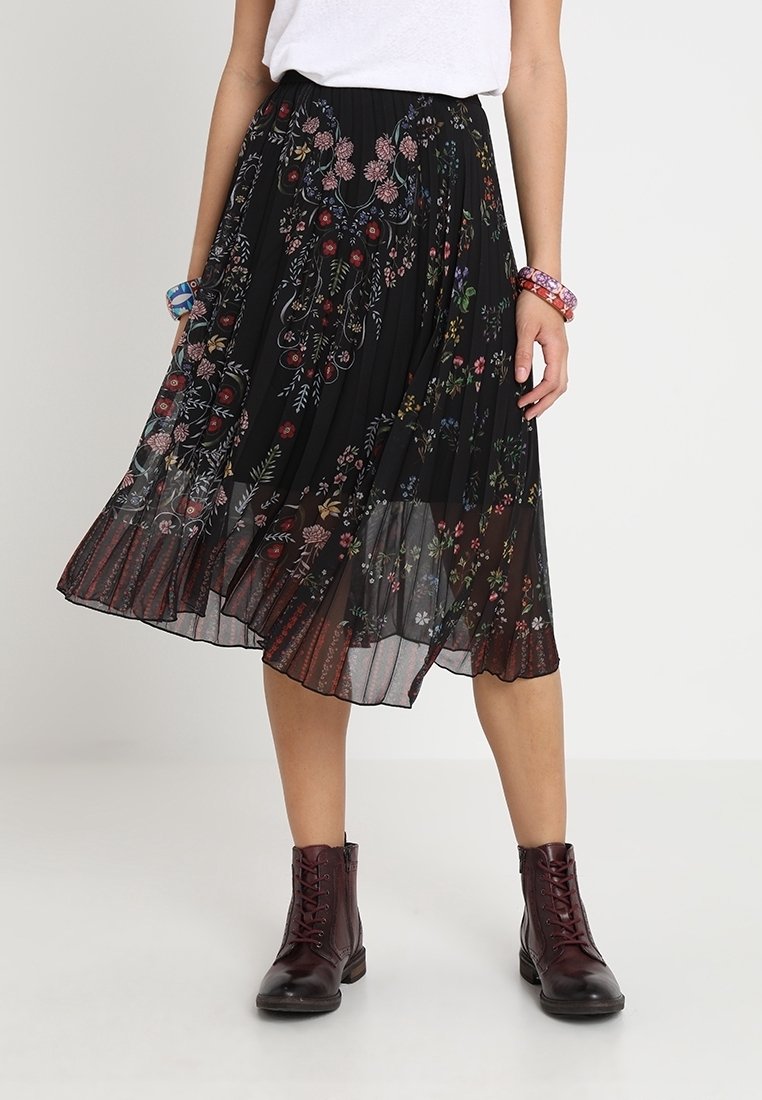 Jupe plissée noire avec broderie florale colorée, tissu transparent et ourlet à rayures horizontales. Associée à des bottines bordeaux.