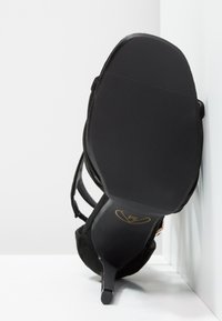 Musta mokkanahkainen korkeakorkoinen sandaali, jossa on terävä kärki, ohut korko ja teksturoitu kumipohja. Sisältää useita koristeellisia hihnoja.