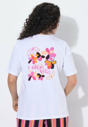 Frau mit lockigem Haar trägt ein weißes T-Shirt mit bunten rosa, orangefarbenen und schwarzen Blumen und dem Schriftzug „dolce vita“ auf dem Rücken.