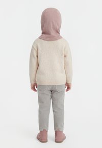 Kind draagt een roze kap, crèmekleurige trui, grijze ribfluwelen broek en roze laarzen, staat met de rug naar voren tegen een witte achtergrond.