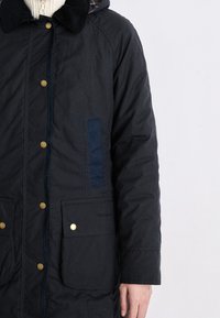 Muž ve tmavě námořnickém kabátu s mosaznými knoflíky, kordovou límcem a velkými předními kapsami, stojí na pozadí jednoduchého bílého pozadí.