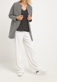 Cappotto lungo a motivi in bianco e nero su un top scuro, abbinato a pantaloni bianchi a gamba larga e sneakers bianche con un accento testurizzato.