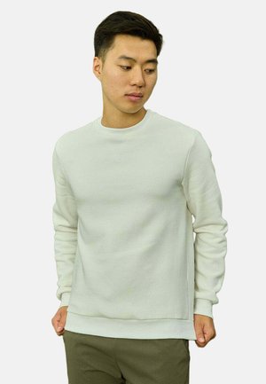 Jeune homme portant un pull blanc uni à col rond et un pantalon olive, regardant vers le bas et sur sa gauche, les mains dans les poches.