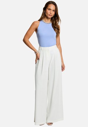 Femme aux cheveux longs portant un haut bleu clair sans manches et un pantalon blanc taille haute à jambes larges, posant avec une main dans la poche.