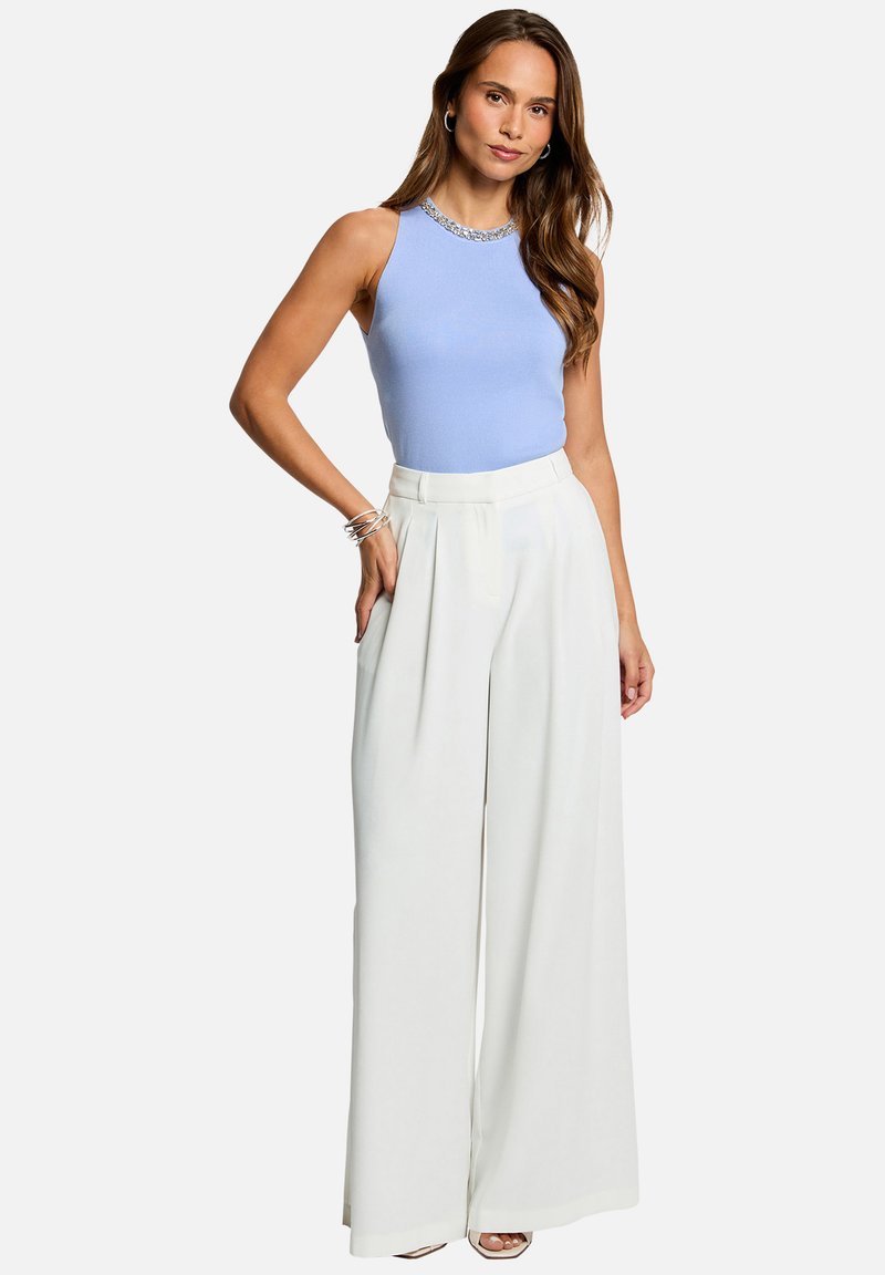 Mujer con pelo largo que lleva una camiseta sin mangas azul claro y pantalones blancos de talle alto y pierna ancha, posando con una mano en el bolsillo.