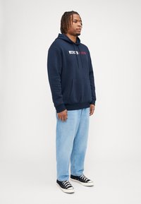 Felpa con cappuccio blu navy, tasca frontale, cappuccio con cordino e dettagli del logo. Abbinata a jeans larghi blu chiaro e sneakers nere.