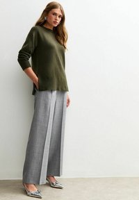 Maglione verde oliva con maniche lunghe, abbinato a pantaloni grigi a pieghe a vita alta e scarpe decorate con dettagli argentati. Texture lisce, vestibilità rilassata.