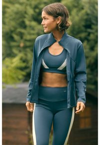 Conjunto atlético azul que incluye un top ajustado y corto con acentos blancos, leggings de cintura alta y una chaqueta a juego con cremallera.