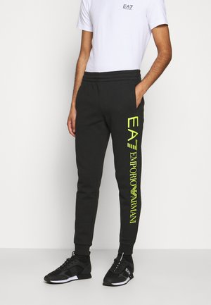 Homme portant un t-shirt blanc et un jogging noir avec le logo jaune néon "EA7 Emporio Armani" sur la jambe droite et des baskets noires.