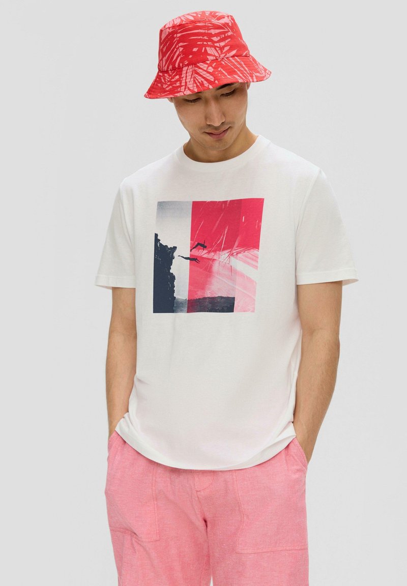 Witte T-shirt met een rechthoekige grafische print in roze en marineblauw, gecombineerd met lichtroze shorts en een rode gebloemde bucket hat.