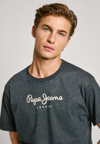 Grå kortermet t-skjorte laget av et mykt stoff. Har "Pepe Jeans London" i hvit kursiv tekst på forsiden. Rund krage.