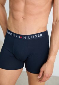Torsos masculino vestido com cuecas boxer Tommy Hilfiger azul-marinho, com o logótipo na cintura visível e mão relaxada ao lado.