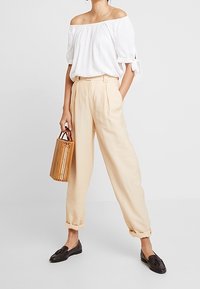 Top bianco scollato con maniche a sbuffo, pantaloni beige a pieghe e mocassini neri. Gli accessori includono una piccola borsa di bambù intrecciato.