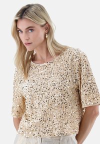Kurzärmeliges Oberteil aus cremefarbenem Stoff, verziert mit goldenen und silbernen Pailletten. Mit rundem Ausschnitt und leicht lockerer Passform.