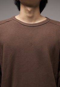 Pull marron en maille gaufrée avec un col rond, arborant un petit logo embossé sur la poitrine et des accents côtelés sur les manches.