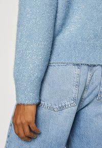 Hellblaues, flauschiges Sweatshirt mit metallischen Fadenakzenten und geripptem Saum, kombiniert mit hochgeschnittenen hellblauen Jeans. Hand ruht auf dem Oberschenkel.