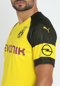 Gul fotbollströja med V-ringning och svarta strukturerade ärmar. Har lila sponsorlogotyp, klubbmärke och Puma-logotyp på axeln.