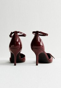 Un paio di scarpe con tacco alto di un brillante rosso scuro con cinturini alla caviglia, viste da dietro su una superficie bianca e uno sfondo chiaro e semplice.