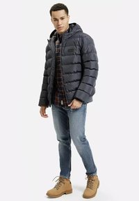 Ciemnoszara kurtka puchowa z poziomymi przeszyciami, z przymocowanym kapturem i zamkiem błyskawicznym z przodu. Noszona na koszuli w kratę i niebieskich dżinsach, z beżowymi butami.