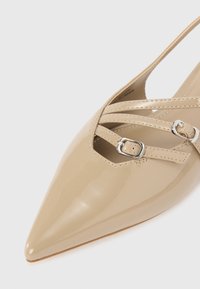 Beige patent leather slingback shoe with pointed toe, featuring cross straps and silver buckles. Smooth, glossy texture with a delicate design.