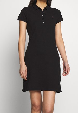 Frau trägt ein schwarzes Polo-Kleid mit kurzen Ärmeln, Kragen und fünf Knöpfen, steht vor einem schlichten hellen Hintergrund.