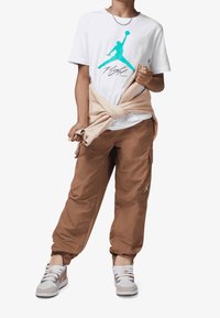 Youth wearing white graphic t-shirt, brown cargo pants, beige jacket tied around waist, and white and beige sneakers, standing against white background.
