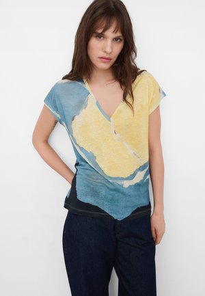 Jonge vrouw met lang bruin haar die een V-hals top met korte mouwen draagt met blauwe, gele en zwarte abstracte patronen en donkerblauwe broek.