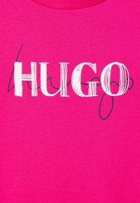 Jasnoróżowy materiał z dużym napisem "HUGO" w białych, stylizowanych literach, nałożonym na delikatne czarne kursywy.