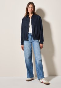 Navyblauwe button-up jas met elastische manchetten, gedragen over een witte top. Combinatie met lichtblauwe wijde jeans en beige sneakers met rode accenten.