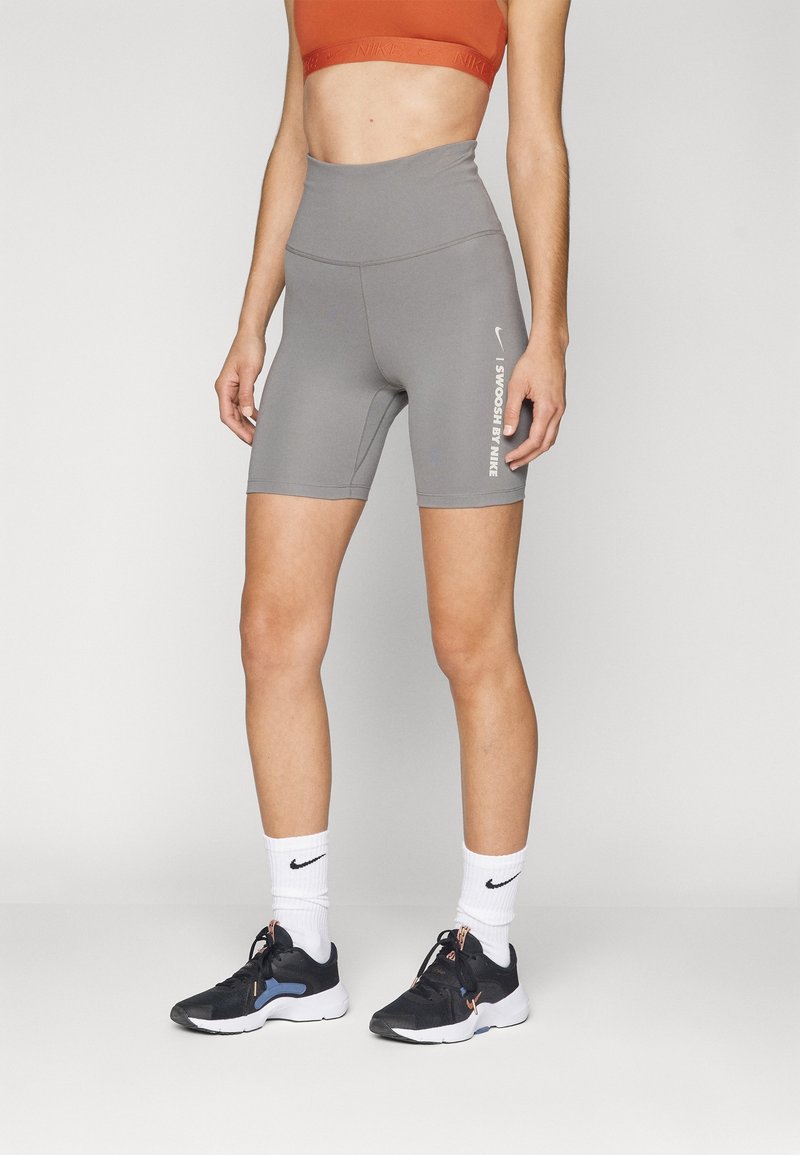 Cuissards de vélo gris taille haute avec une texture lisse et le logo blanc "Swoosh by Nike" sur le côté, associés à des chaussures de sport noires et des chaussettes blanches.