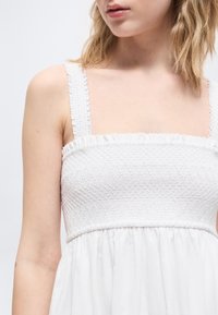 Γυναίκα με λευκό φόρεμα χωρίς μανίκια, ελαστικό μπούστο με σφηκοφωλιά και βολάν στα τιράντες, εμφανίζεται από τους ώμους έως το μέσο του κορμού πάνω σε απλό φόντο.