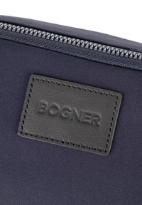 Temno modra tkaninska torbica z črno usnjeno etiketo z vtisnjenim napisom "BOGNER." Na zgornji robni strani je srebrna zadrga. Enostavna, ploska zasnova.