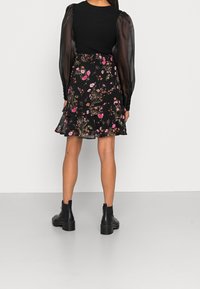 Jupe fleurie noire avec motifs roses et verts, ourlet à volants superposés, associée à un haut côtelé noir et des bottines noires. Détails de manches longues transparentes.