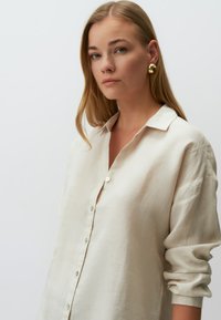 Lichtbeige linnen shirt met een knoopsluiting aan de voorkant, kraag en opgerolde mouwen. De stof heeft een natuurlijke textuur en een subtiele glans.
