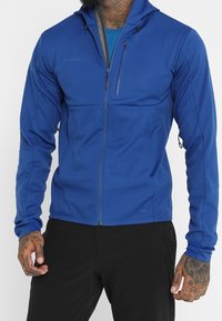 Giacca blu a conchiglia morbida con chiusura a zip frontale, cappuccio e due tasche laterali. Presenta una vestibilità slim con elementi regolabili e tessuto strutturato.