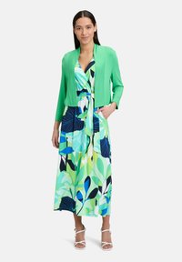 Cardigan vert sur une robe maxi à fleurs multicolores présentant des motifs de feuilles bleues, vertes et noires. Design ouvert et élégant, longueur genou.