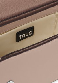 Bolso de cuero texturizado rosa con ribete marrón, interior beige y una etiqueta negra que dice "TOUS". Detalles en hardware plateado visibles.