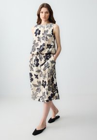 Beige twee-delige outfit met zwarte bloemenpatronen, bestaande uit een mouwloze top en een midi-rok met zakken. Zwart instapschoenen.