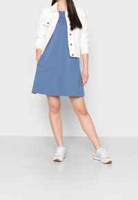 Niebieska sukienka bez rękawów z bocznymi kieszeniami, w połączeniu z białą kurtką dżinsową. Model nosi jasne sneakersy z niebieskimi i różowymi akcentami.