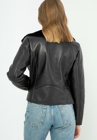 Giubbotto di pelle nera con un design aderente e cuciture sulle spalle pronunciate, dotato di un collare di pelliccia nera e una superficie liscia. Indossato sopra jeans in denim.