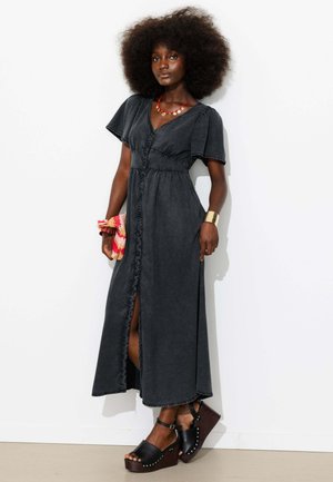 Femme aux grands cheveux bouclés portant une robe longue noire boutonnée sur le devant, des sandales à plateforme noires, un collier rouge, un bracelet manchette doré et tenant une pochette colorée.