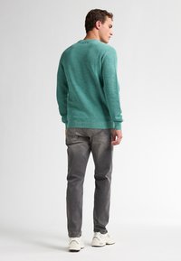 Turquoise gestructureerde gebreide trui, grijze broek en witte sportschoenen. Zichtbare naaddetails op de broek en de manchetten. Achteraanzicht.