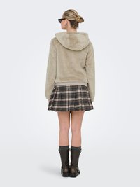 Hooded, beige fur jacket with a plush texture, paired with a plaid, pleated skirt. Model wears black knee-high boots and glasses.